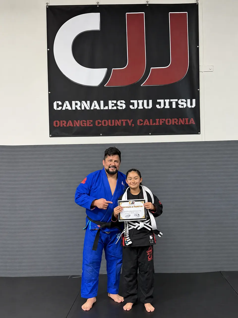 Professor Rudy of Carnales Jiu Jitsu with a student holding her promotion certificate